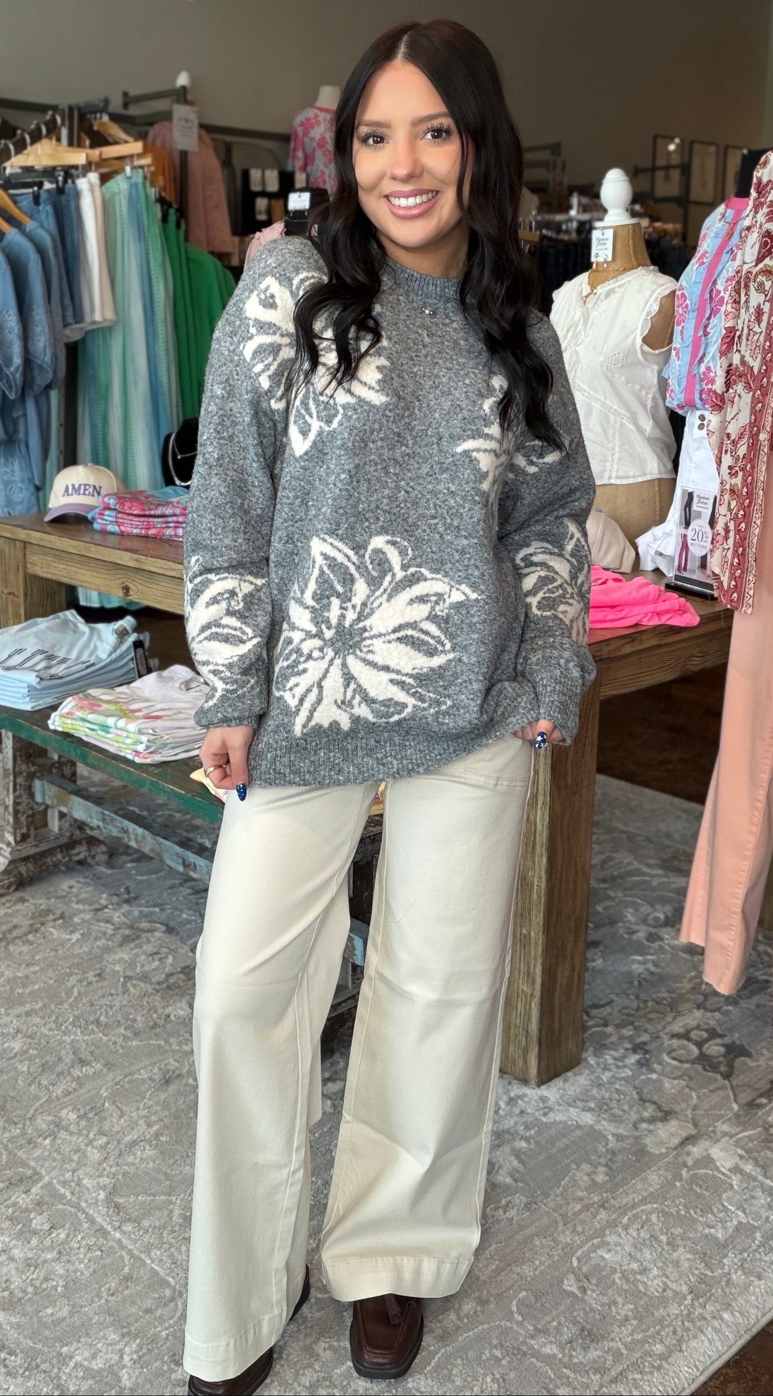 A woman stands in a clothing boutique wearing a gray knit sweater with large white floral patterns and wide-leg cream pants. Clothing racks, mannequins, and store fixtures are visible in the background.