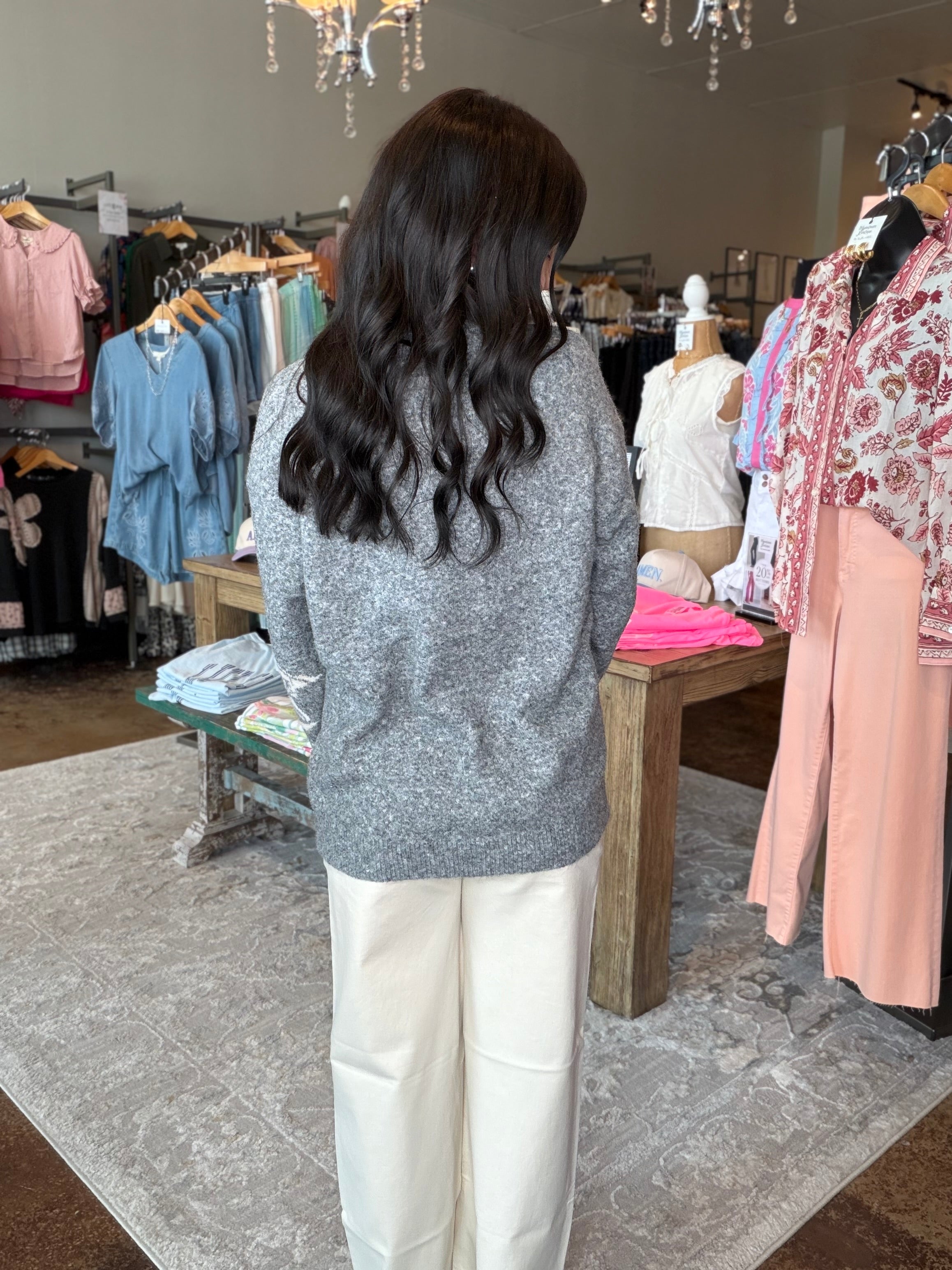 A woman with long, wavy dark hair is seen from behind, wearing a gray knit sweater and cream wide-leg trousers inside a clothing boutique. Clothing racks, a wooden table, and a patterned rug are visible in the background.
