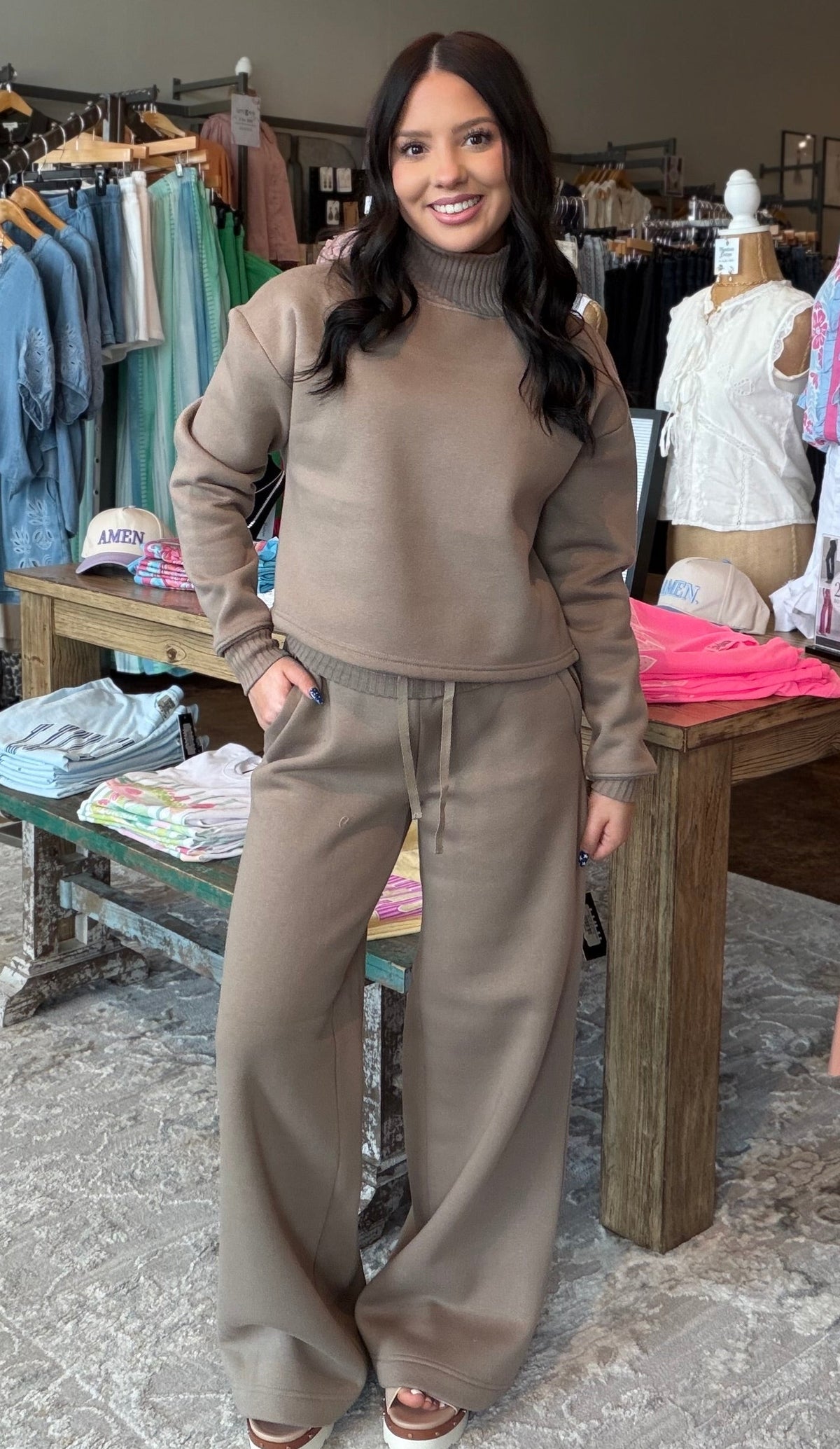 A woman modeling a matching taupe brown lounge set consisting of a turtleneck top and wide-leg sweatpants, standing inside a clothing boutique.