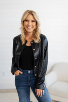 Woman wearing a black leather jacket with gold buttons, standing against a white wall.