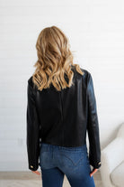 Person wearing a black leather jacket and blue jeans against a white wall.