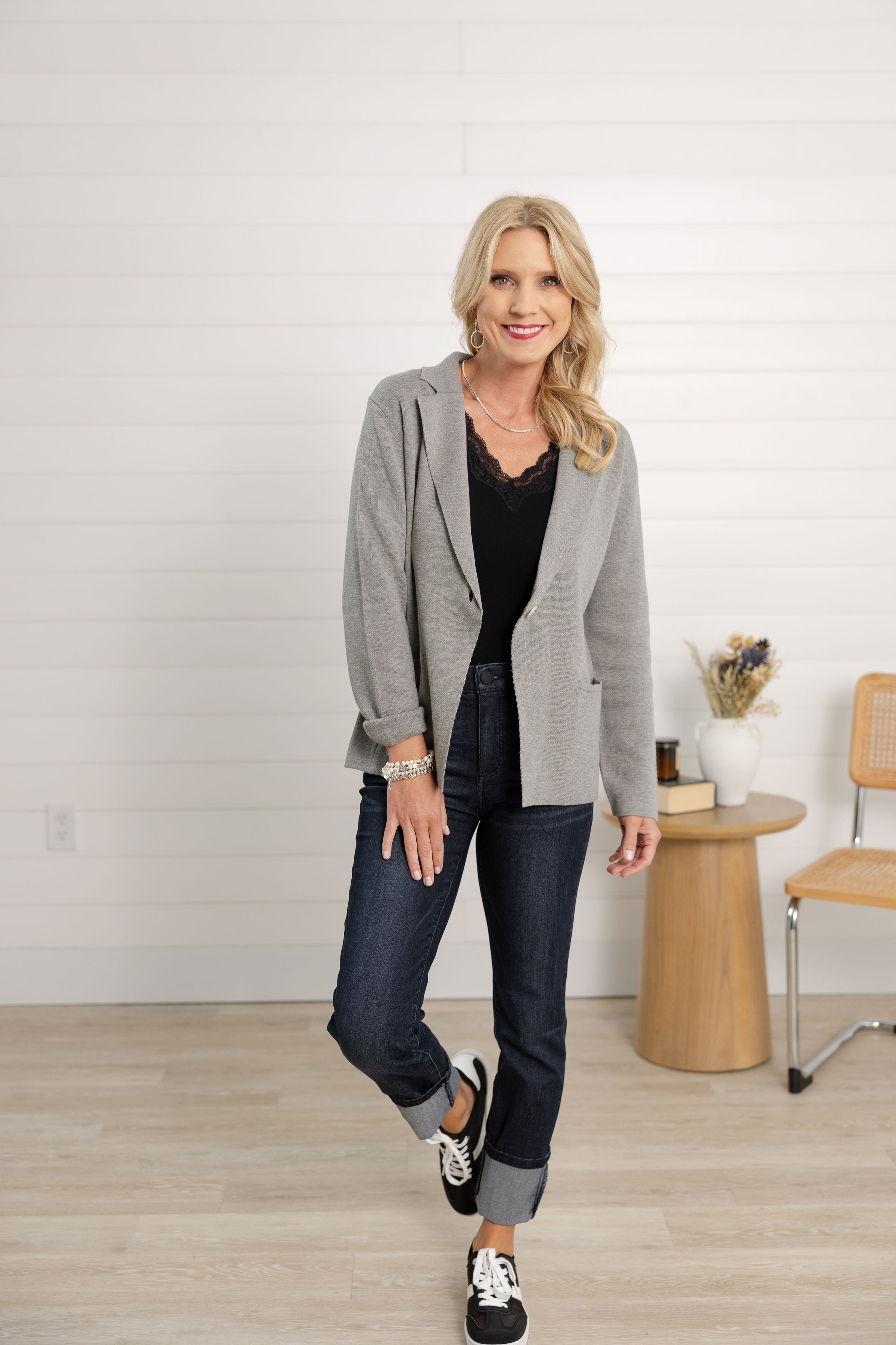 Woman wearing a gray blazer, black top, and jeans in a room with a wooden table and chair.