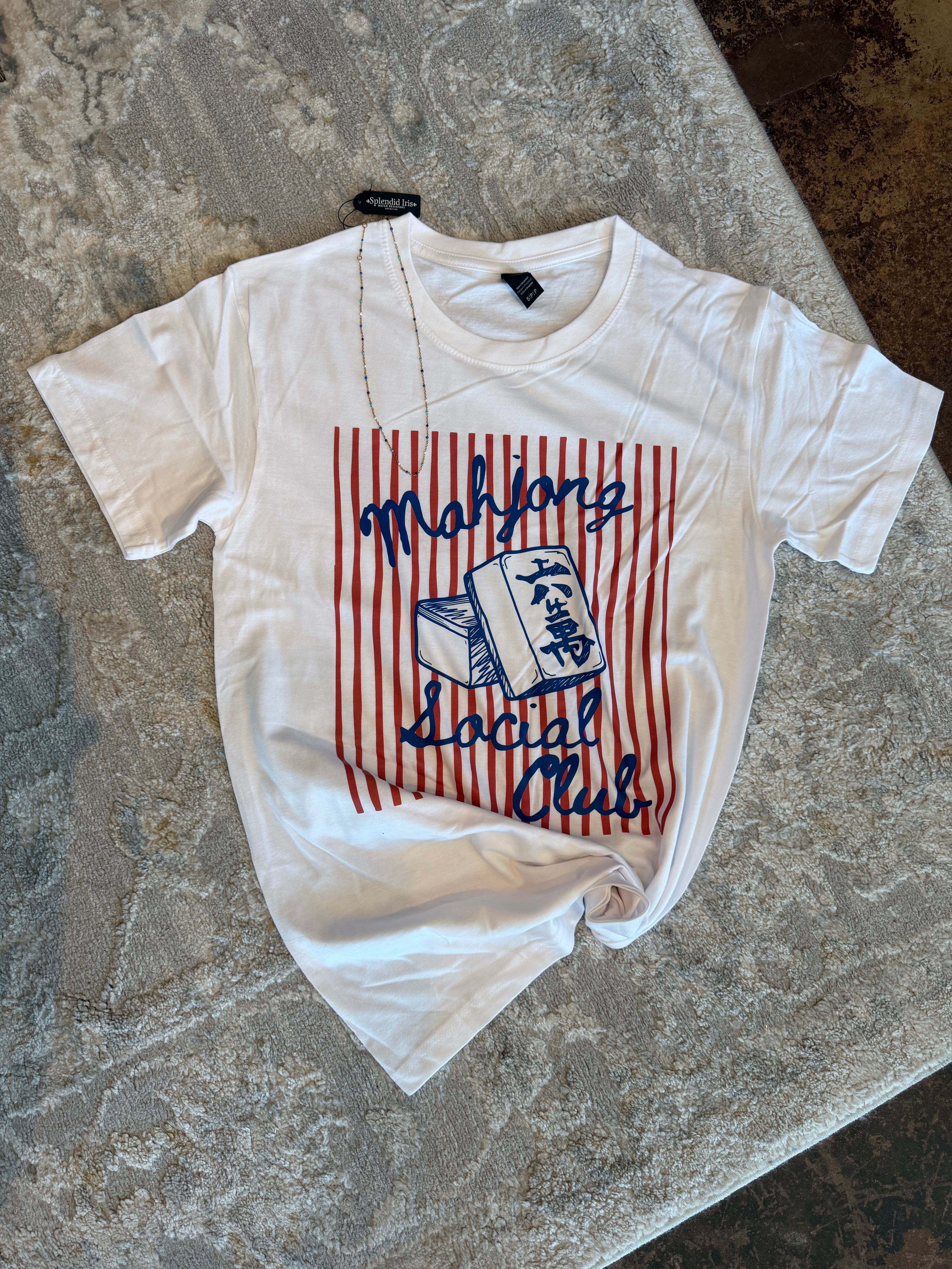 A white short-sleeve graphic T-shirt laid flat on a textured gray surface. The shirt features a red and white vertical stripe graphic with blue text reading 'Majiang Social Club' and a blue book illustration.