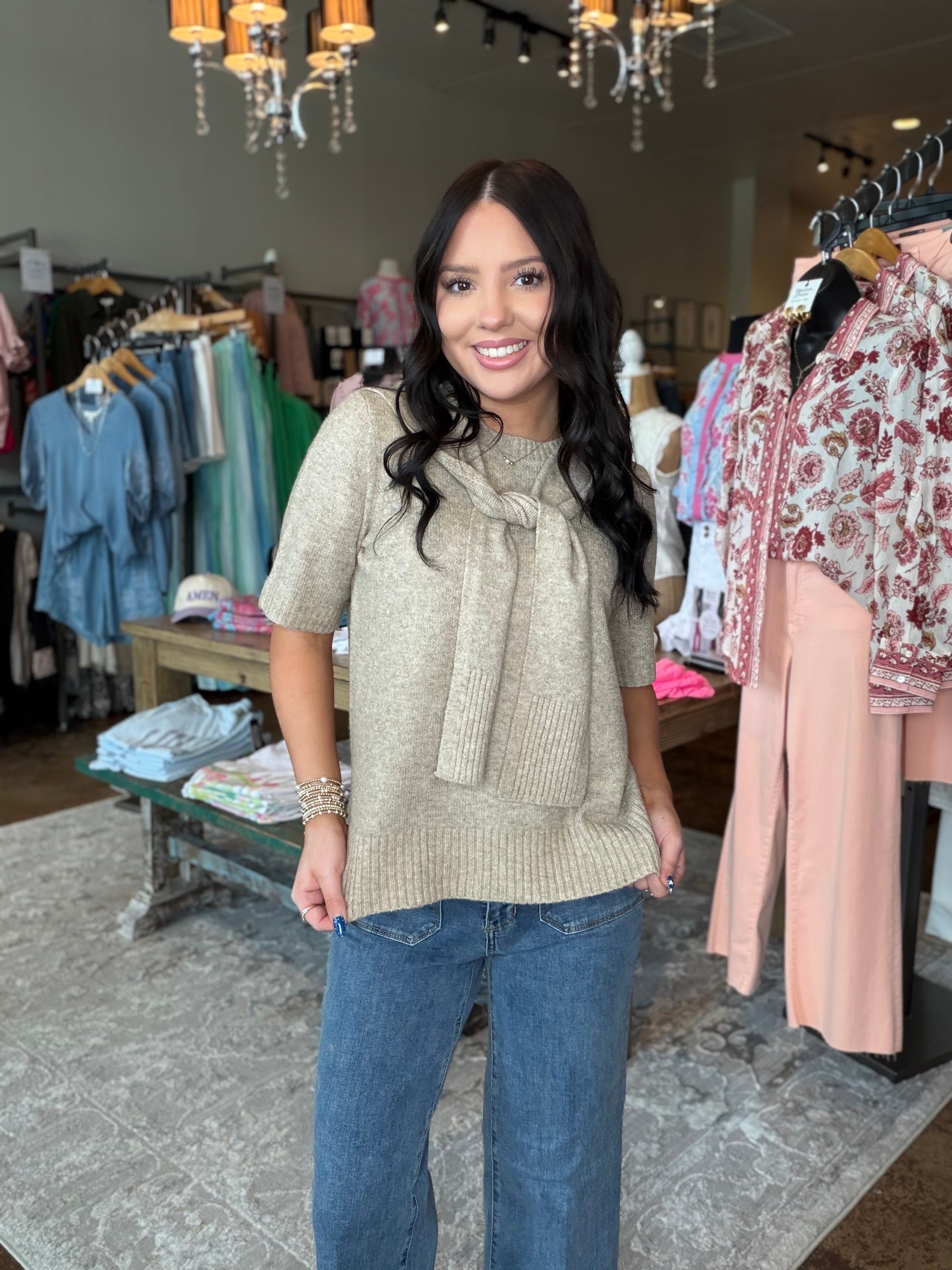 A woman modeling a beige short-sleeve knit top with a large front bow detail, paired with blue jeans, standing inside a clothing boutique.