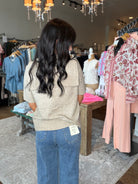 A woman with long, wavy dark hair is seen from behind in a clothing boutique, wearing a beige short-sleeve knit top and blue jeans. Clothing racks with pink and floral tops are visible on either side of the store interior.