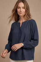 A woman wearing a navy blue long-sleeve blouse with a split neckline and gathered sleeves, posed against a neutral beige background.