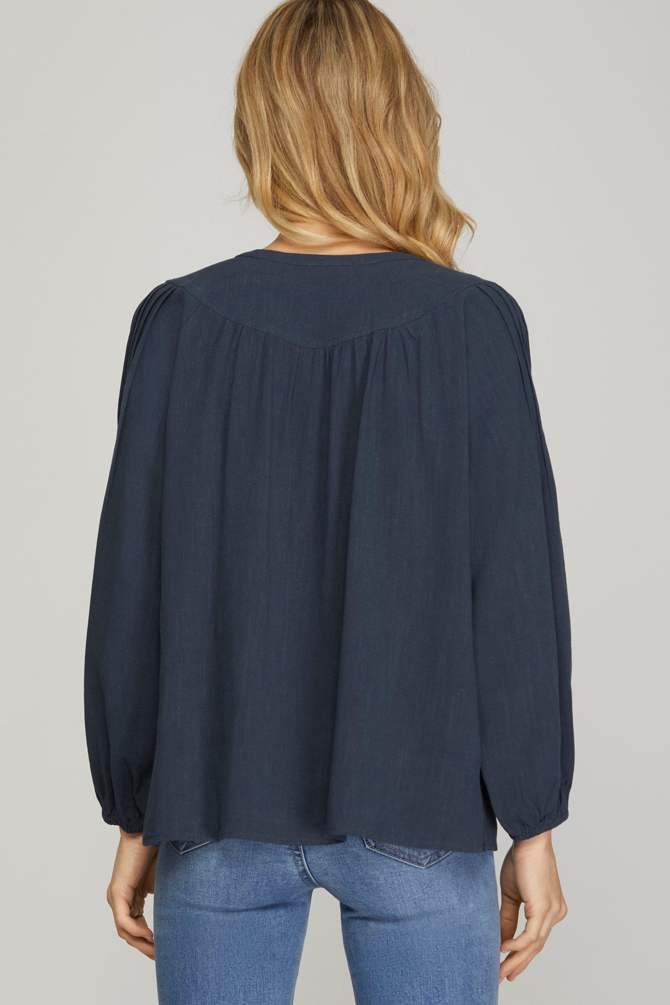 A woman viewed from the back wearing a dark navy blue long-sleeve blouse with gathered shoulders and blue jeans, against a plain light gray studio background.