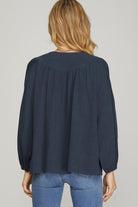 A woman viewed from the back wearing a dark navy blue long-sleeve blouse with gathered shoulders and blue jeans, against a plain light gray studio background.