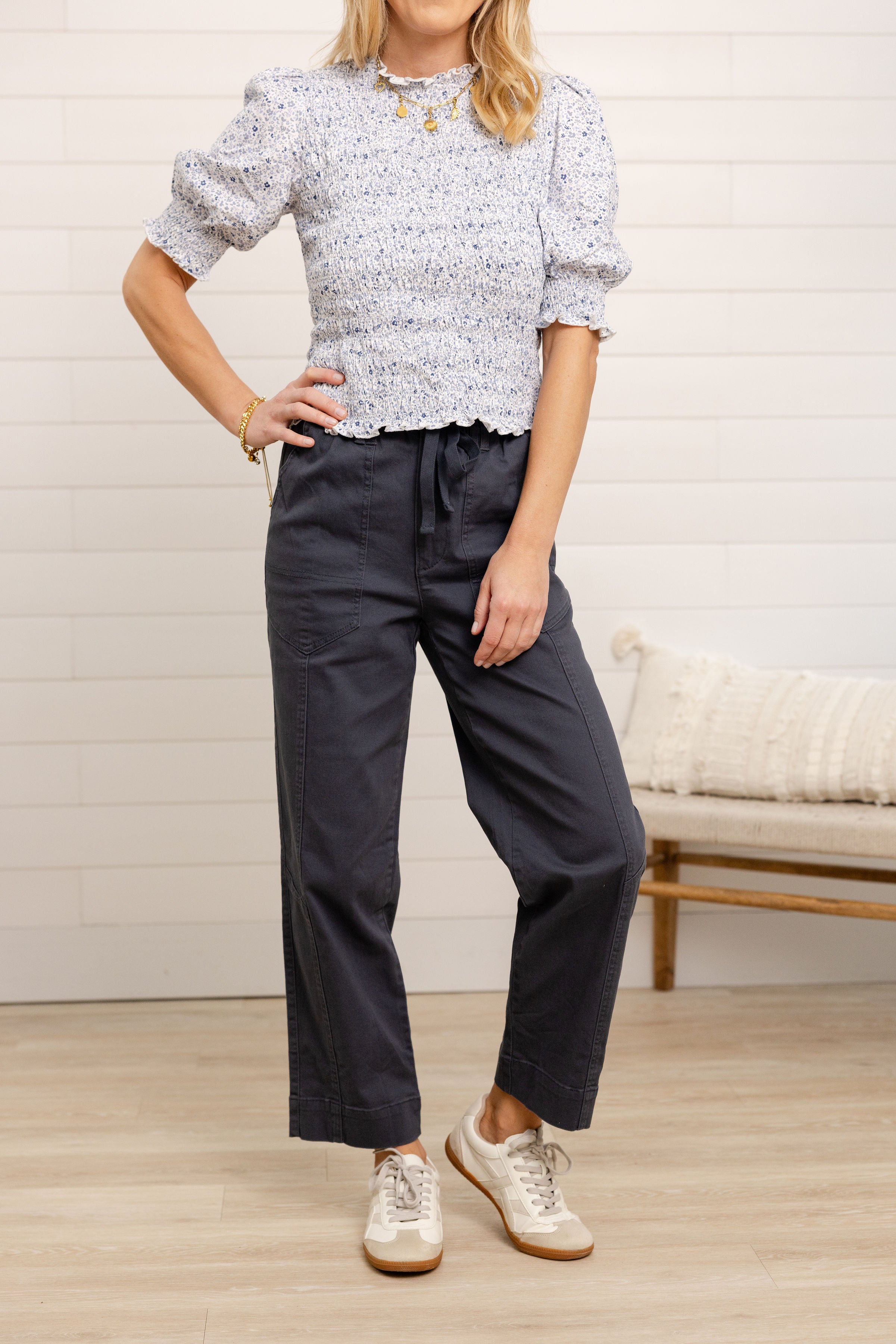 Woman wearing a floral blouse and dark pants standing in a room with a light-colored wall and wooden floor.