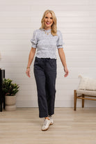 Woman wearing a patterned blouse and dark pants in a room with a white wall and wooden floor.
