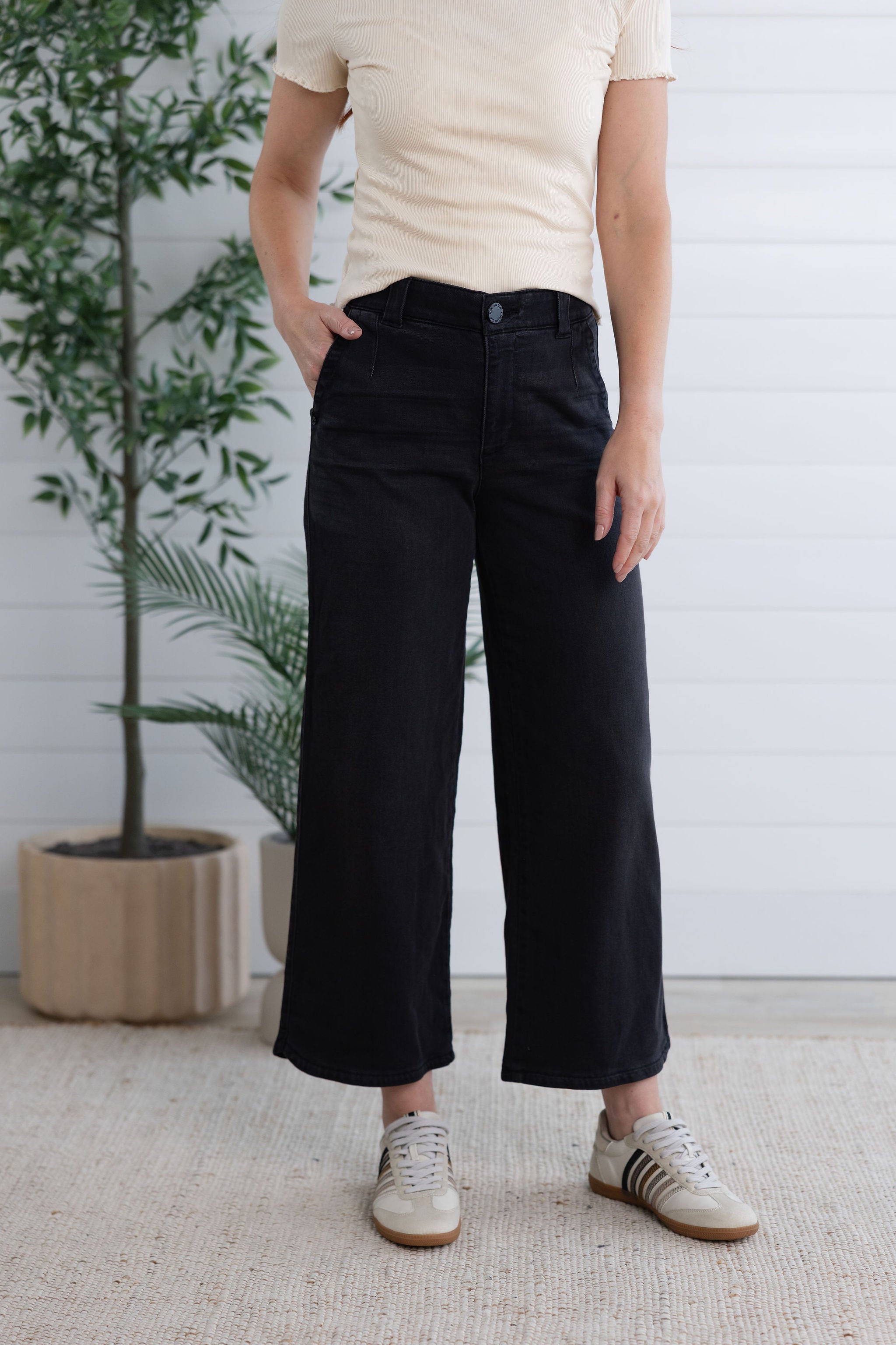 Person wearing black pants and a beige shirt standing indoors with a plant in the background