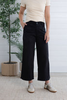 Person wearing black pants and a beige shirt standing indoors with a plant in the background