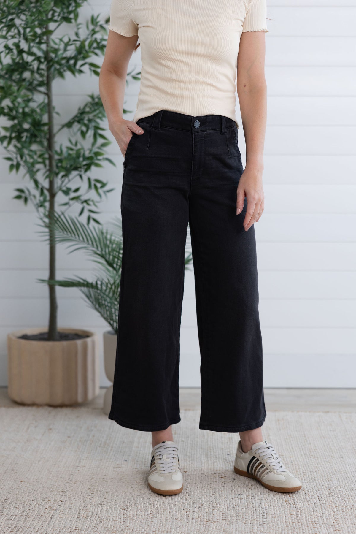 Person wearing black pants and a beige shirt standing indoors with a plant in the background