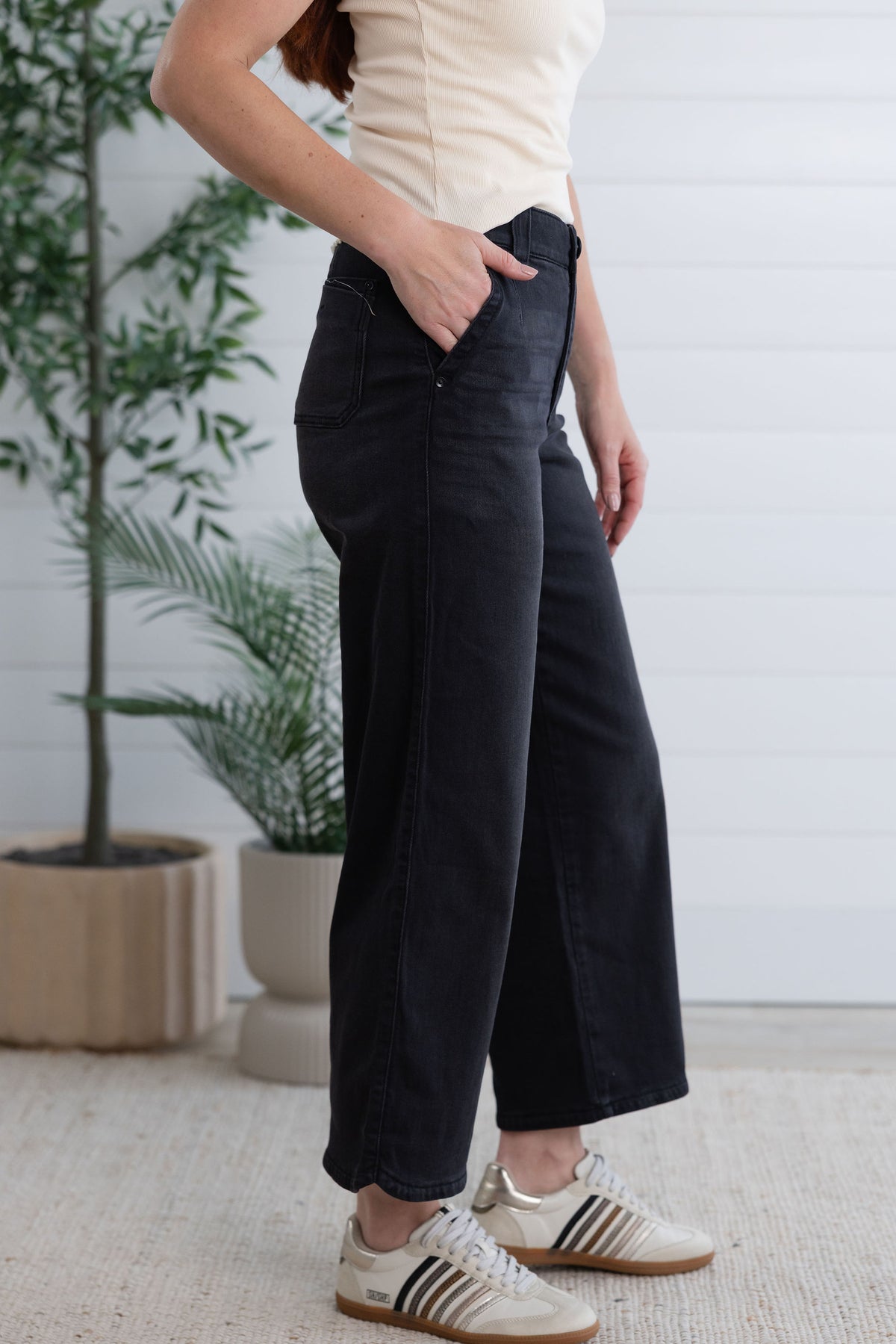 A person wearing black wide-leg cropped jeans, a cream ribbed top, and white Adidas sneakers, standing in a bright indoor setting with potted plants and a white paneled wall.
