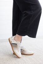A person wearing off-white Adidas Samba sneakers with black and beige stripes, paired with black wide-leg pants, standing on a light textured rug.