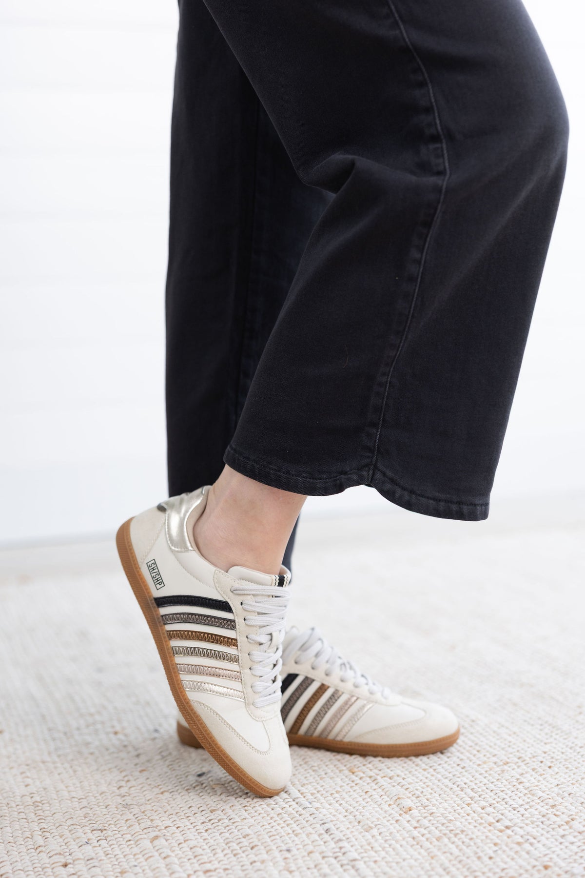 A person wearing off-white Adidas Samba sneakers with black and beige stripes, paired with black wide-leg pants, standing on a light textured rug.