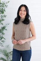 Woman wearing a brown sleeveless knitted sweater against a white background with green plants.