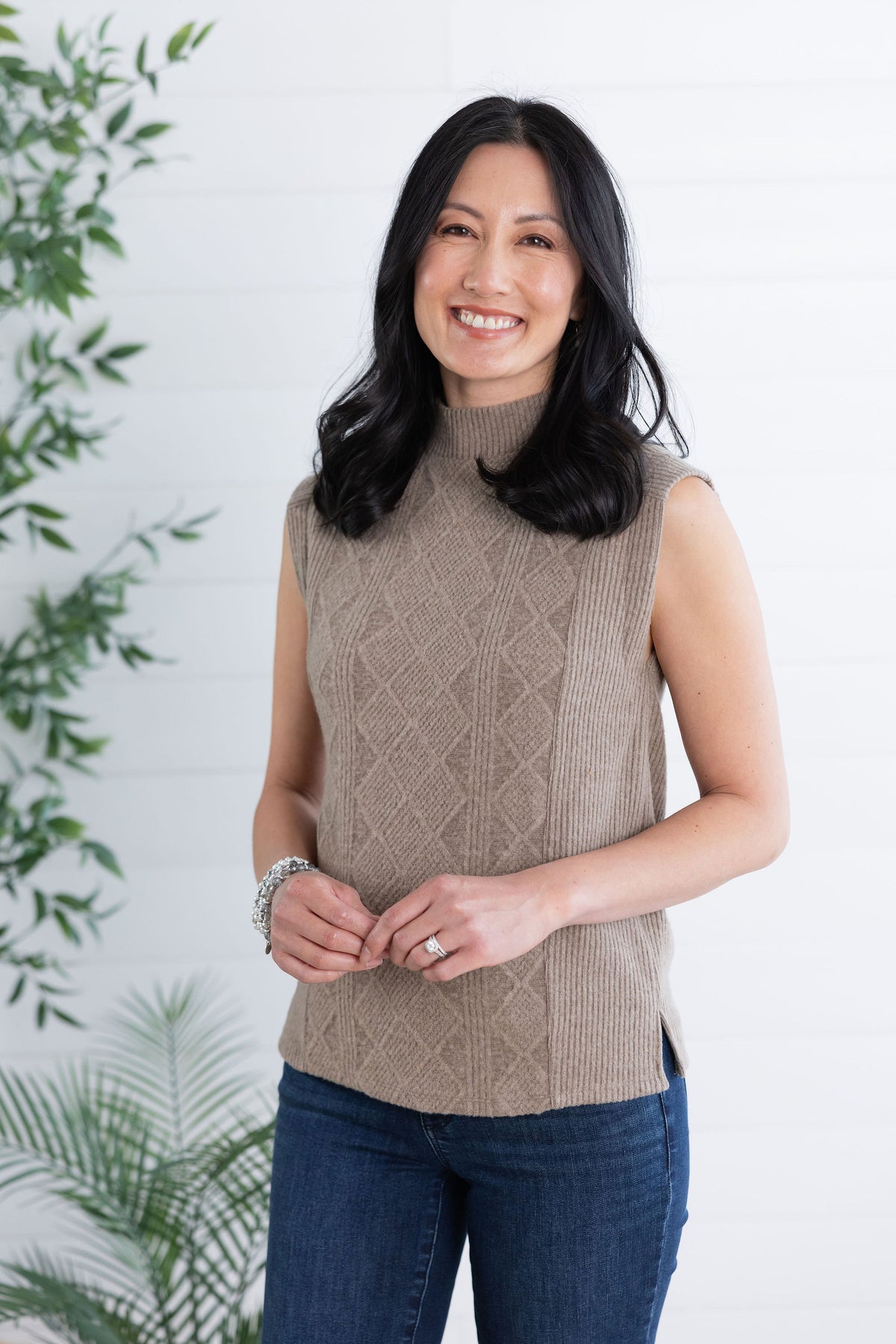 Woman wearing a brown sleeveless knitted sweater against a white background with green plants.