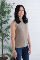 Woman wearing a beige sleeveless sweater and blue jeans standing in front of a white wall with a plant.