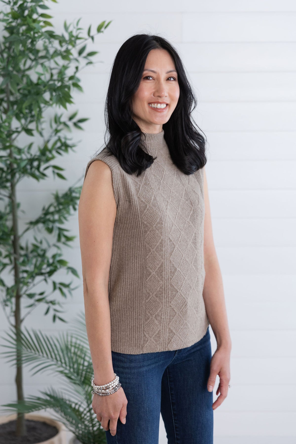 Woman wearing a beige sleeveless sweater and blue jeans standing in front of a white wall with a plant.