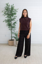 A woman stands indoors wearing a sleeveless dark plum knit top and dark plaid wide-leg pants, with black flats. She is posed in front of a white paneled wall with a potted tree and rug.