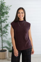 A woman wearing a sleeveless, mock-neck knit top in a deep plum purple color, paired with dark plaid pants, standing in front of a white paneled wall with potted plants.