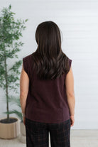 A woman viewed from behind wearing a sleeveless burgundy knit top and dark plaid pants, standing in front of a white paneled wall with a potted plant to the left.