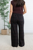 A woman shown from the back wearing dark plaid wide-leg trousers and a sleeveless burgundy top, standing indoors against a white paneled wall with potted plants.