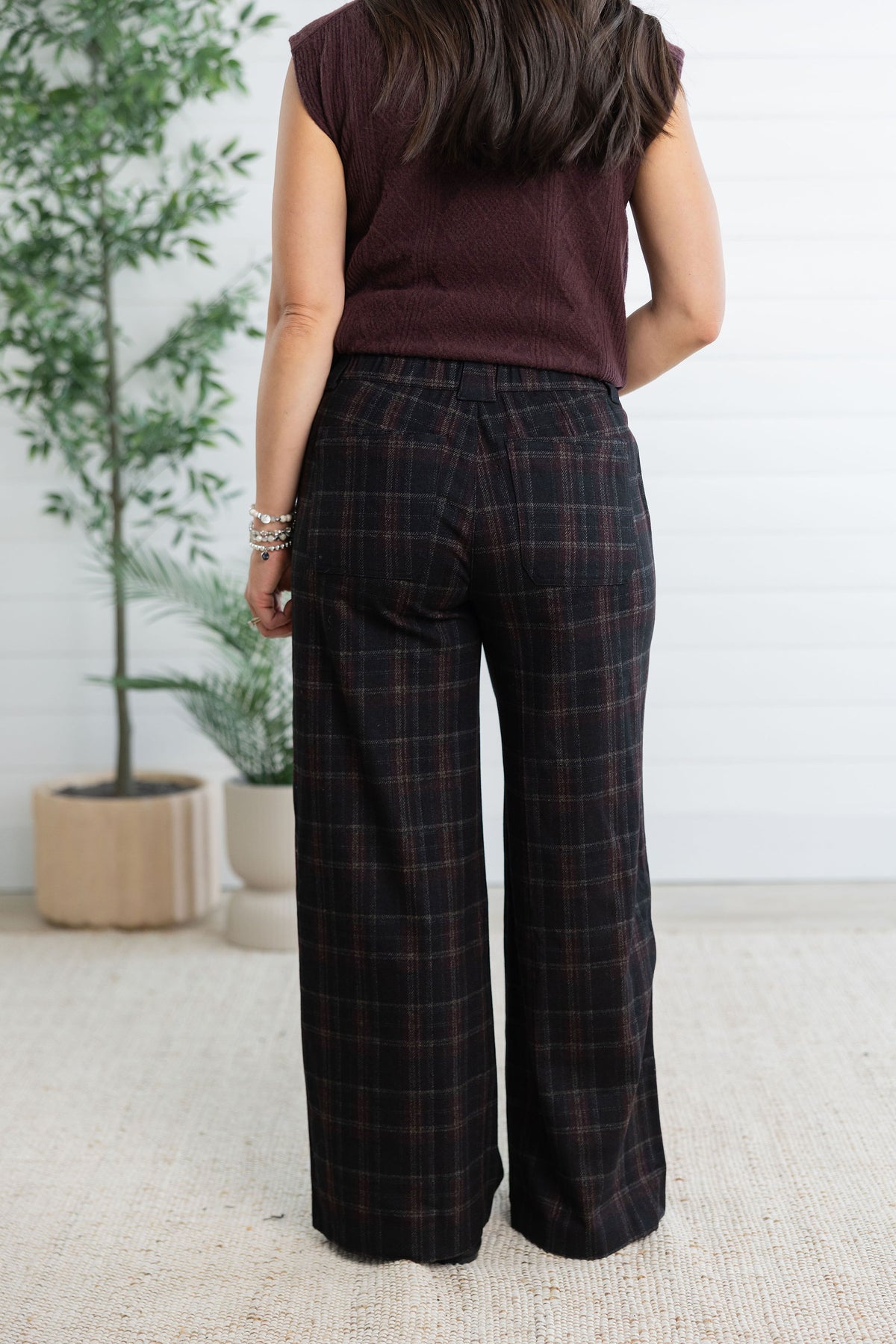 A woman shown from the back wearing dark plaid wide-leg trousers and a sleeveless burgundy top, standing indoors against a white paneled wall with potted plants.