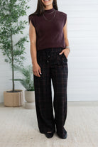 A woman wearing a sleeveless dark plum knit top and dark plaid wide-leg pants, standing in a bright indoor setting with potted plants and a white paneled wall.
