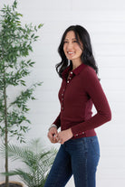 A woman wearing a fitted long-sleeve button-front collared top in deep burgundy and blue jeans, standing indoors against a white paneled wall with green plants visible on both sides.