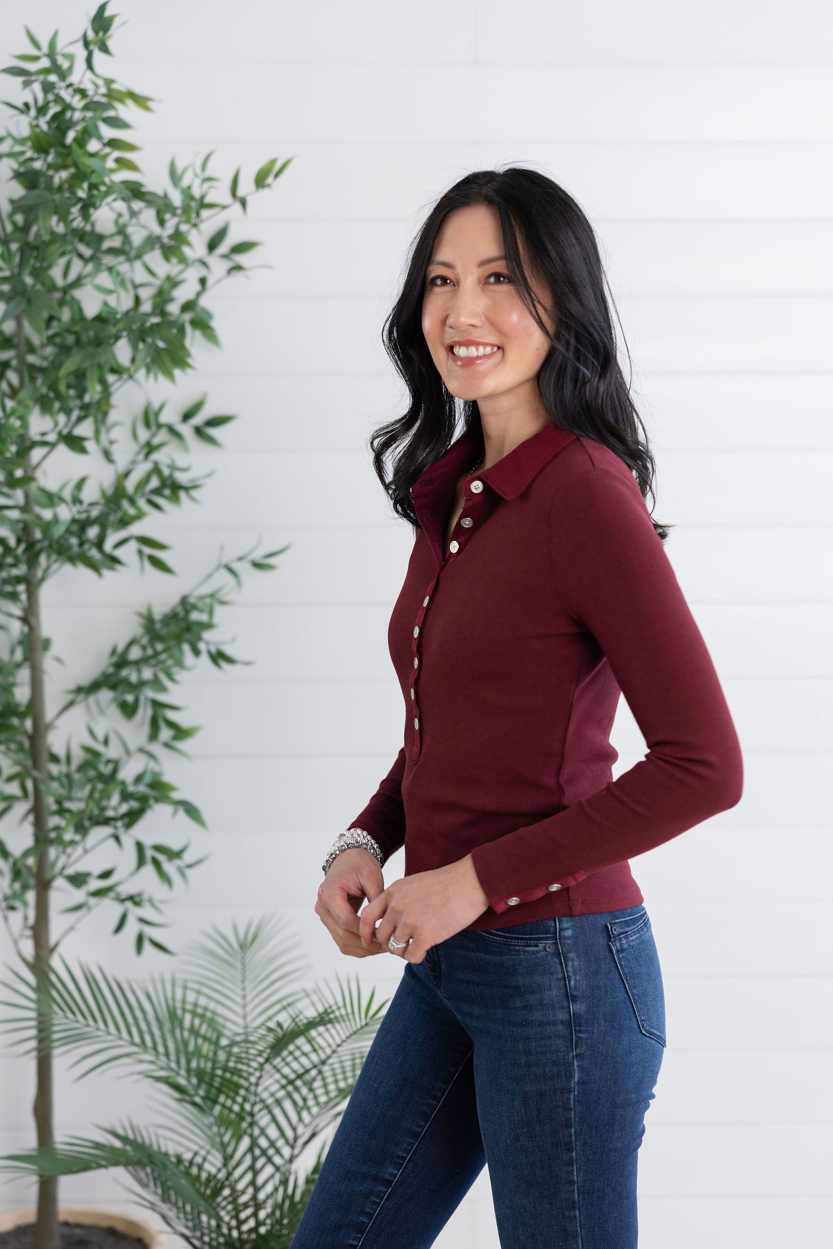 A woman wearing a fitted long-sleeve button-front collared top in deep burgundy and blue jeans, standing indoors against a white paneled wall with green plants visible on both sides.