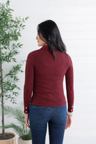 A woman viewed from the back wearing a fitted long-sleeve mock neck top in burgundy and blue jeans, standing against a white paneled wall with potted plants on the left.