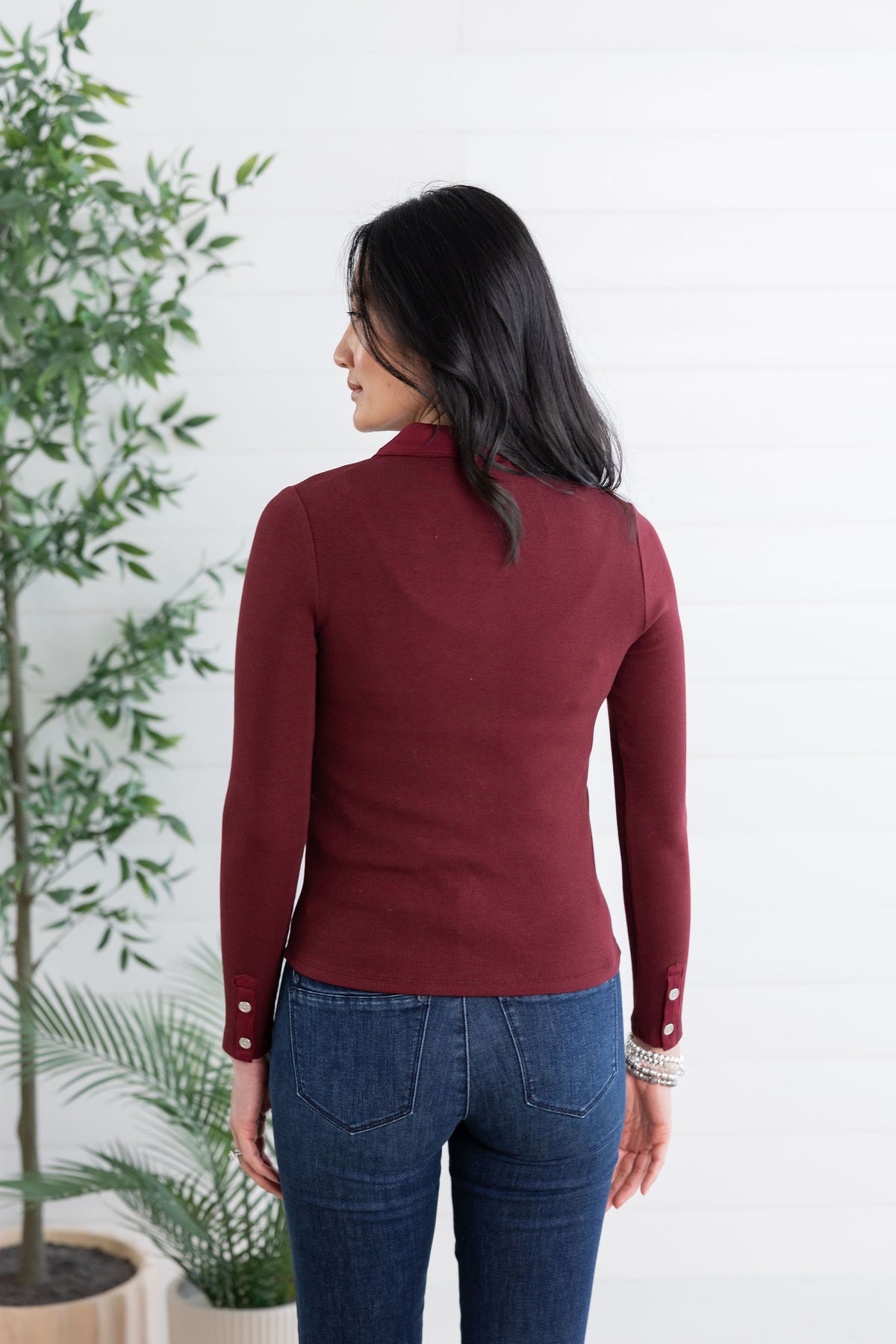 A woman viewed from the back wearing a fitted long-sleeve mock neck top in burgundy and blue jeans, standing against a white paneled wall with potted plants on the left.