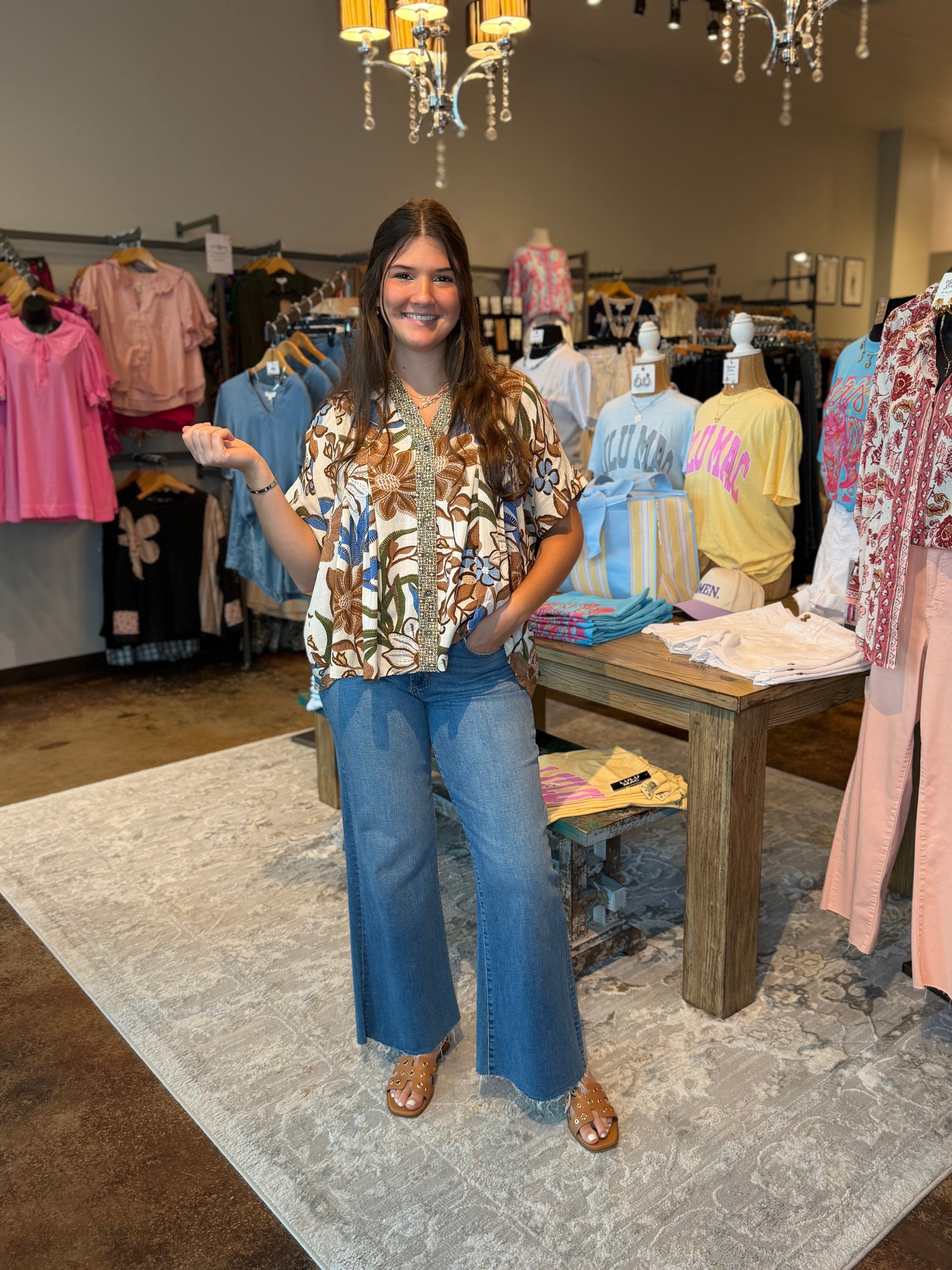 A woman stands in a clothing boutique wearing a brown and beige botanical print short-sleeve button-up shirt with blue wide-leg jeans and brown sandals. Clothing racks, mannequins, a wooden table, and hanging chandeliers are visible in the background.