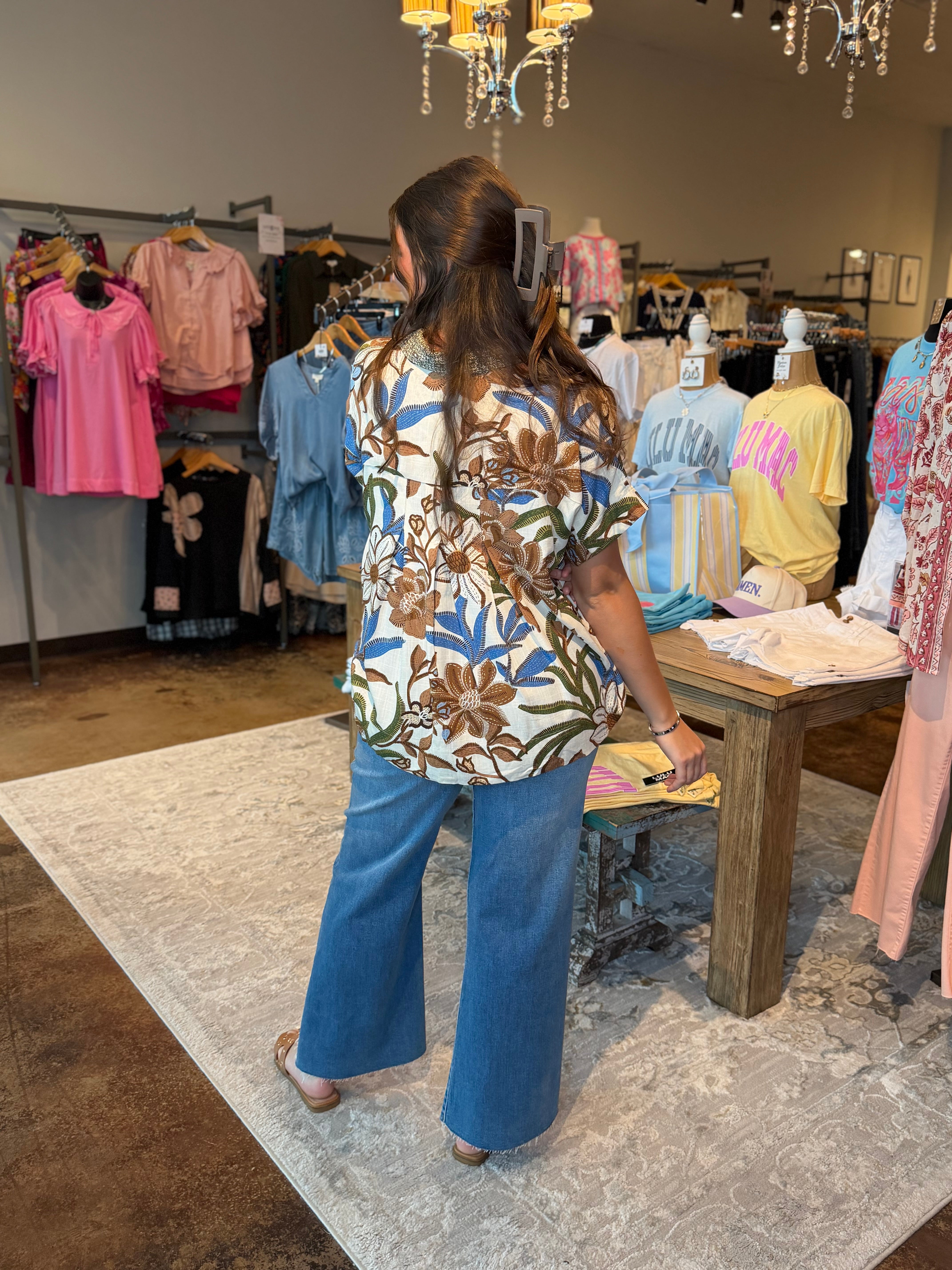 A woman stands in a clothing boutique wearing a short-sleeve floral print top and wide-leg blue jeans, viewed from behind. The store has racks of colorful garments, mannequins, a wooden table, and hanging chandeliers.