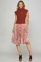 A woman modeling a sleeveless rust-brown mock-neck top paired with a pleated pink and white abstract print midi skirt, styled with black platform sandals against a light gray studio background.