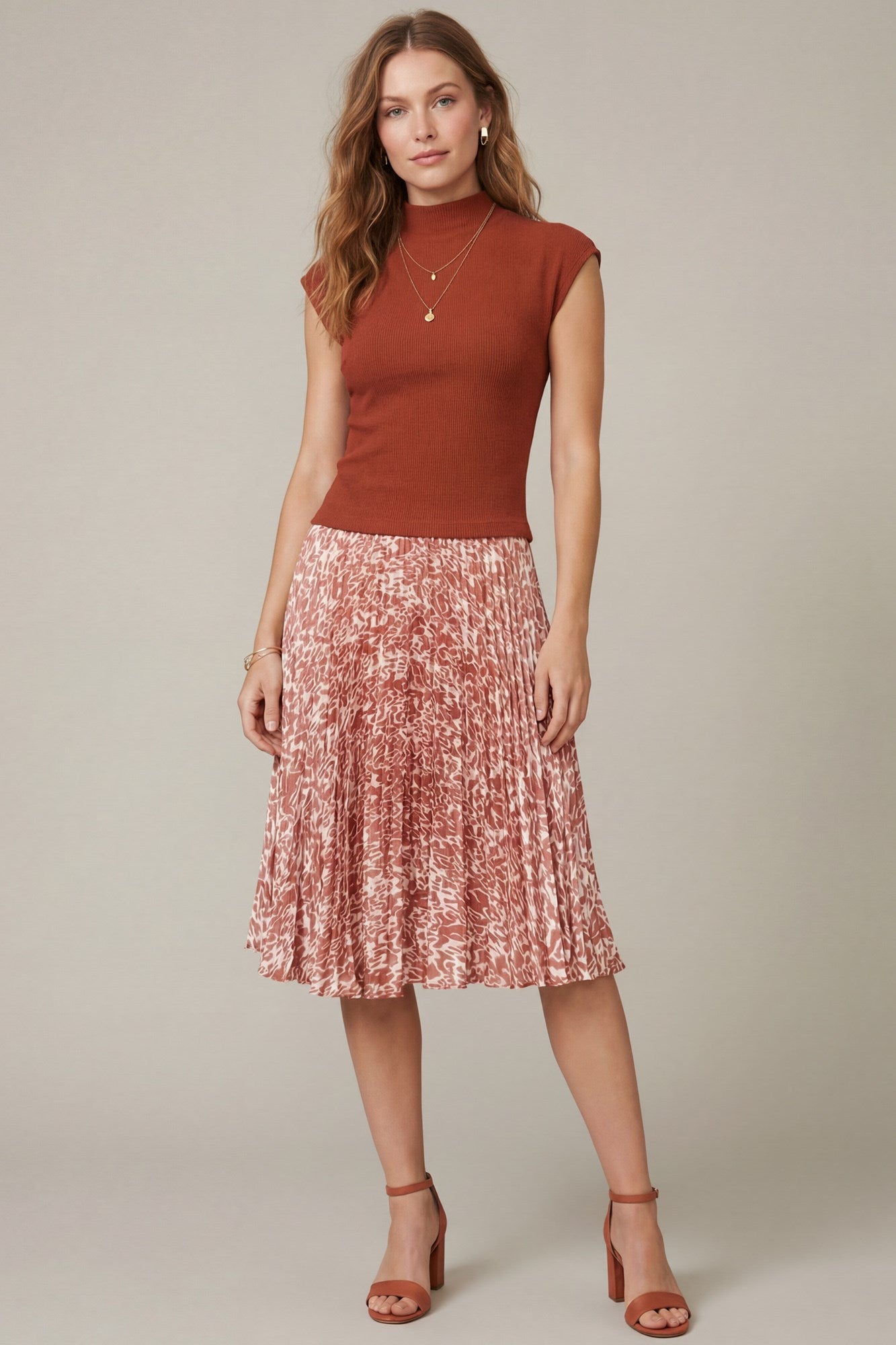 A woman modeling a rust-colored sleeveless mock-neck top paired with a pleated midi skirt in a pink and red abstract pattern, against a solid light gray studio background.