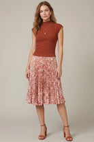 A woman modeling a rust-colored sleeveless mock-neck top paired with a pleated midi skirt in a pink and red abstract pattern, against a solid light gray studio background.