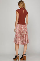 A woman shown from the back wearing a sleeveless rust-brown top and a pleated midi skirt with a pink, red, and white abstract pattern. She is wearing black platform sandals against a light gray studio background.