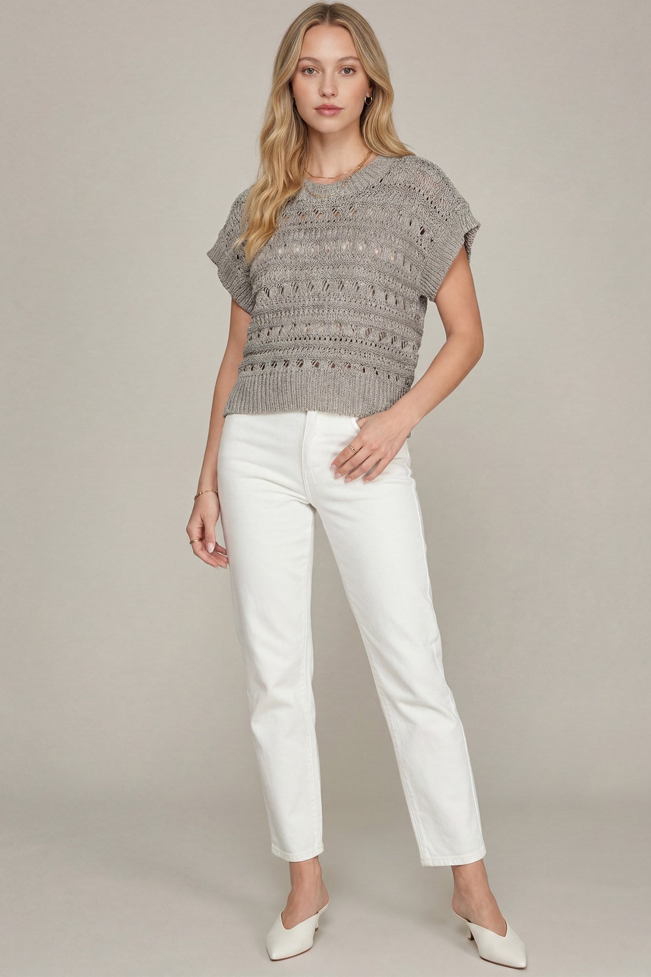 A woman modeling a gray textured short-sleeve knit top with white cropped pants and white pointed-toe heels, standing against a plain light gray studio background.