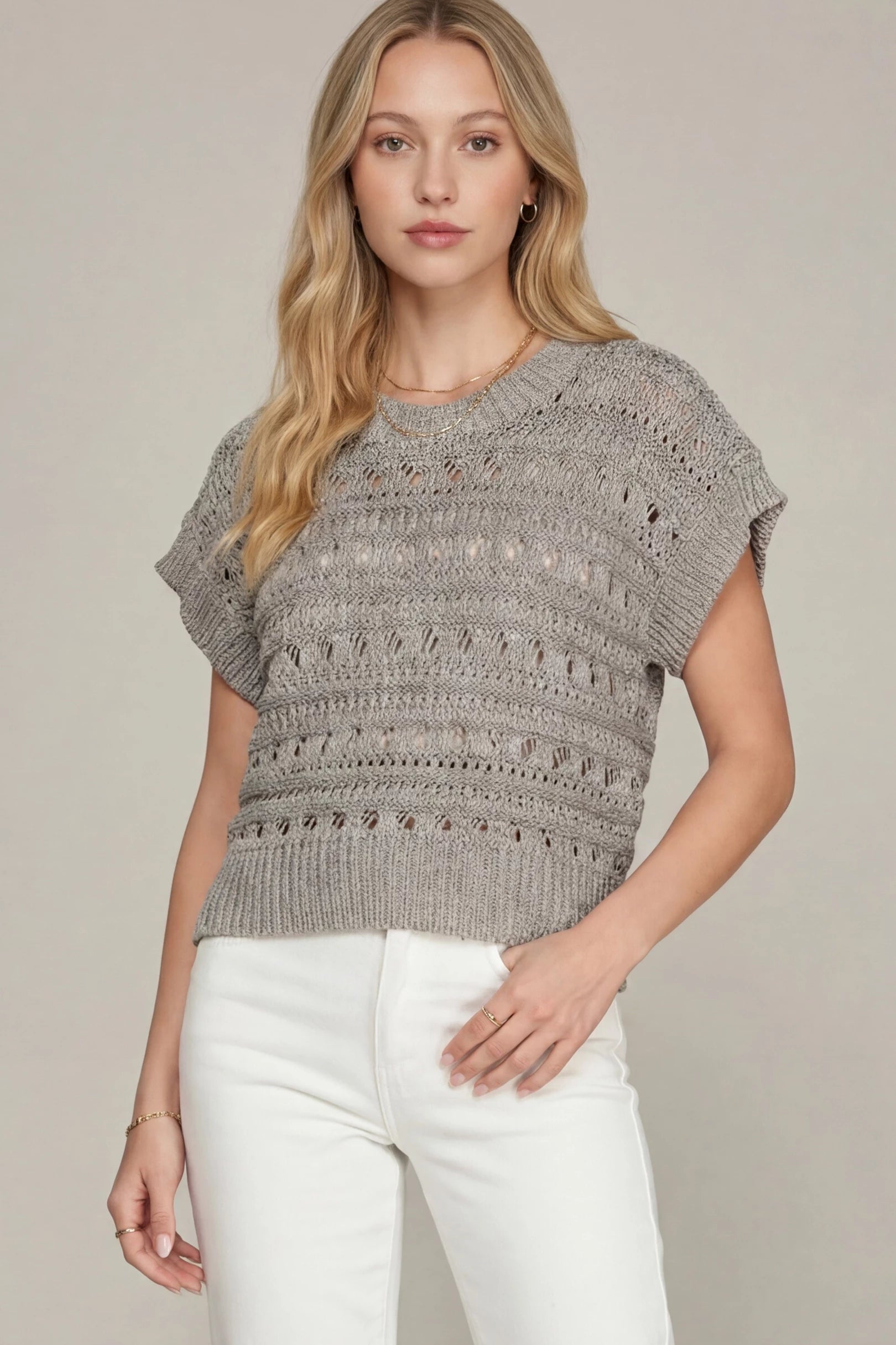 A woman wearing a gray textured knit short-sleeve top with white pants, standing against a plain beige studio background.