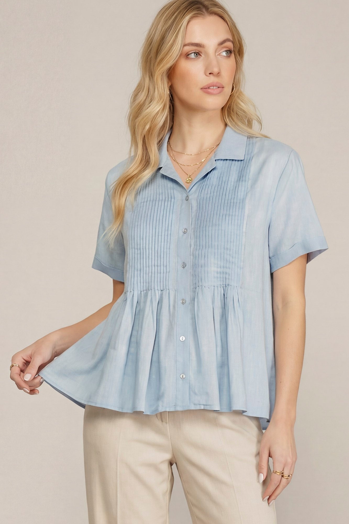 A woman wearing a light blue short-sleeve button-up blouse with a gathered peplum waist and beige pants, standing against a plain beige studio background.