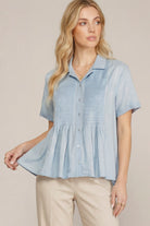 A woman wearing a light blue short-sleeve button-up blouse with a gathered peplum waist and beige pants, standing against a plain beige studio background.