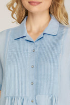 Close-up of a woman wearing a light blue collared shirt dress with vertical pleats and silver buttons.