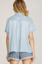 A woman shown from the back wearing a light blue short-sleeve button-up shirt and denim shorts against a plain light beige studio background.