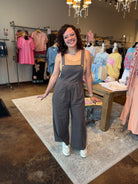 A woman with curly dark hair and glasses stands in a clothing boutique wearing a dark gray wide-leg jumpsuit over a tan tank top and white sneakers. The boutique has racks of colorful clothing, a wooden table, and a light-colored rug.