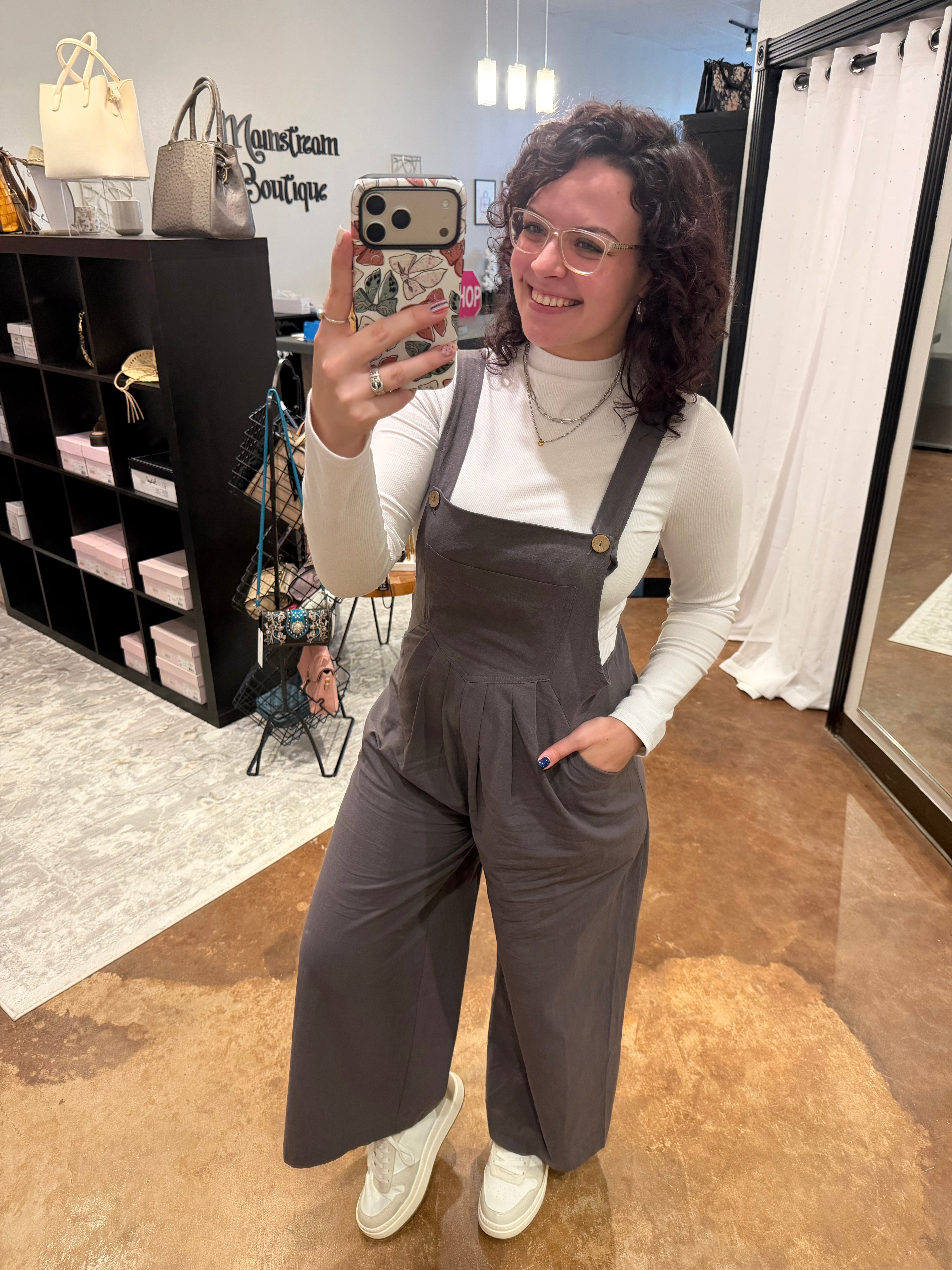 A woman takes a mirror selfie in a boutique wearing dark gray wide-leg overall pants over a white long-sleeve top and white sneakers. The boutique interior features a black shelving unit with handbags and a white curtain in the background.