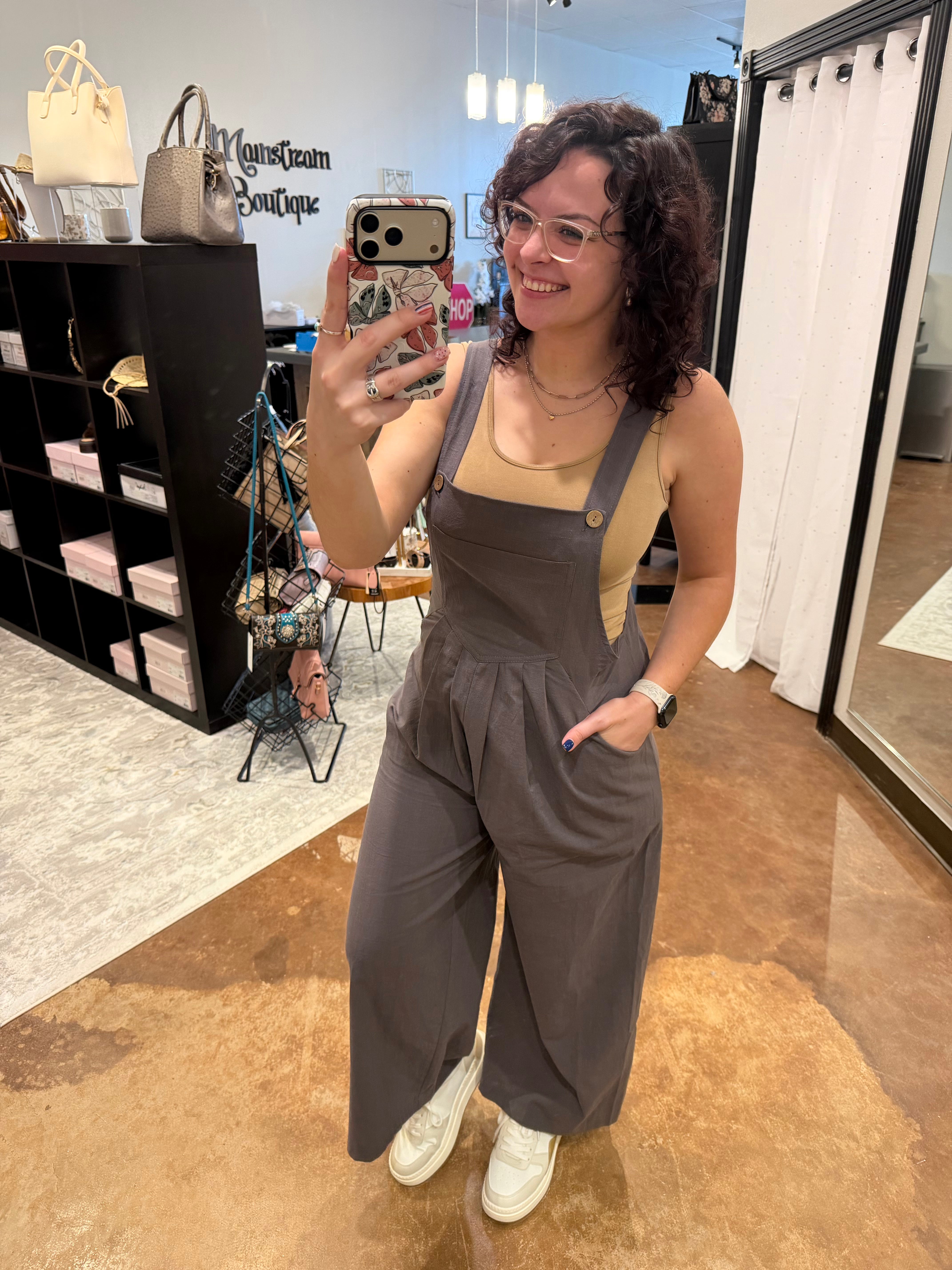 A woman takes a mirror selfie in a boutique wearing dark gray wide-leg overall jumpsuit with a beige tank top underneath and white sneakers. The boutique interior features a brown tiled floor, black shelving, and a white curtain.