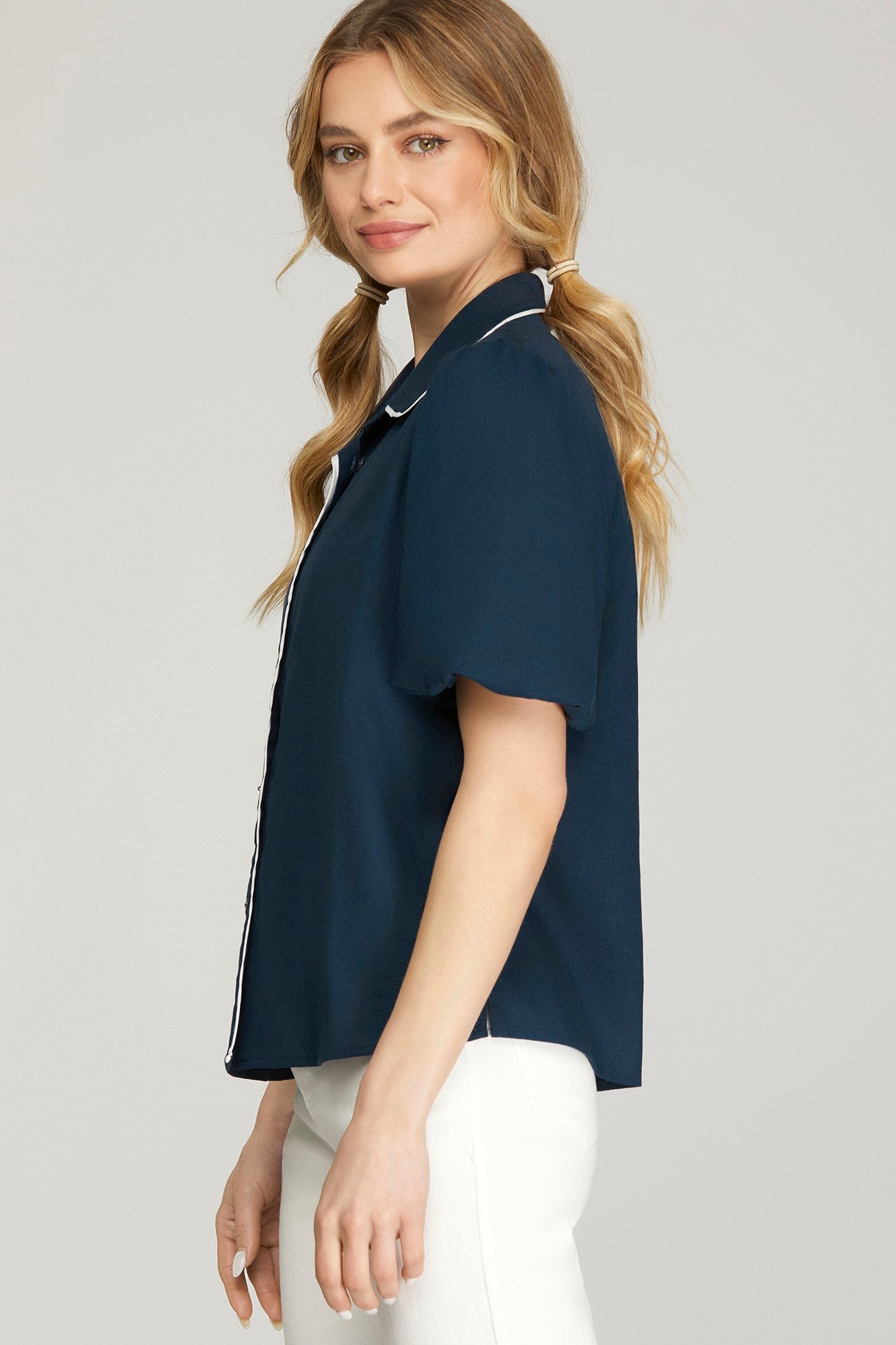 A woman wearing a navy blue short-sleeve button-up shirt with white piping details, paired with white pants, shown from a side-back angle against a light gray studio background.